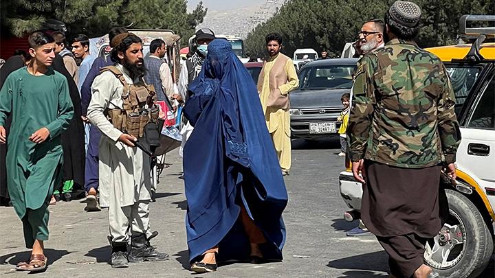 Seorang perempuan yang mengenakan Burqa berjalan melewati Pasukan Taliban yang memblokir jalan-jalan di sekitar bandara, di Kabul, Afghanistan (27/8/2021). Taliban juga melarang perempuan menekuni olahraga karena dinilai tidak sesuai dengan syariat Islam yang diyakini, dengan alasan khawatir bagian tubuh perempuan akan terekspose ketika berolahraga. (Foto: Reuters).